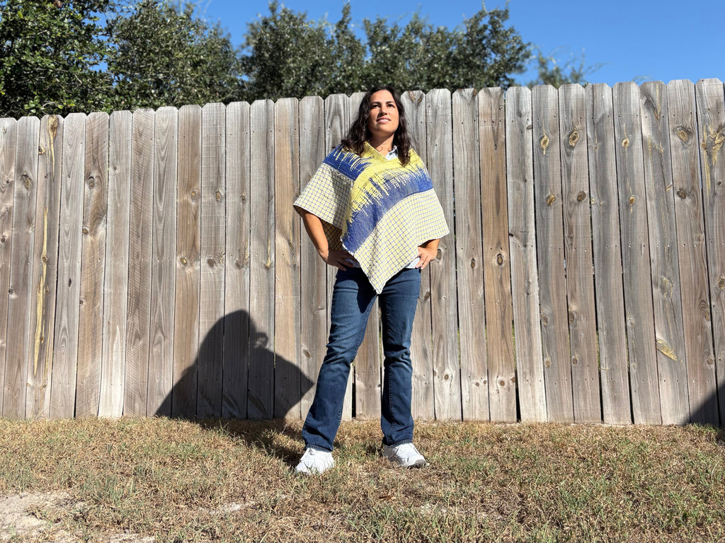 A woman stands outdoors in front of a wooden fence wearing a cream poncho-style top with blue and yellow geometric patterns, paired with blue jeans and white sneakers.