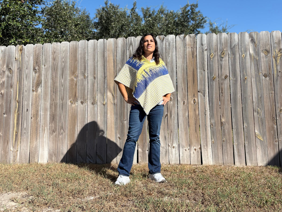 A woman stands outdoors in front of a wooden fence wearing a cream poncho-style top with blue and yellow geometric patterns, paired with blue jeans and white sneakers.